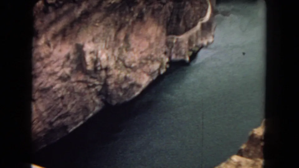 1967 8mm frame showing rugged canyon rock and green-blue river water below, likely near the Hoover Dam area based on clip metadata.