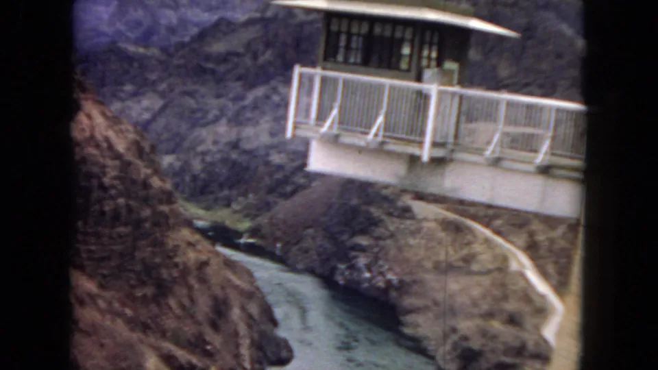 1967 home-movie frame of a white-railed overlook, winding road, and river cutting through rocky desert canyon terrain near Hoover Dam.