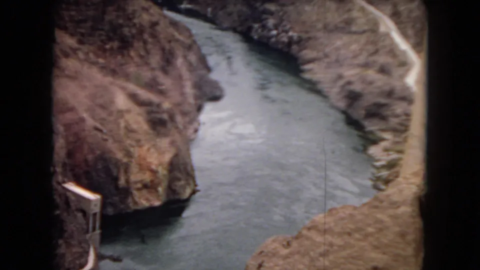 1967 Arizona home-movie frame showing a narrow river below reddish canyon walls with a concrete dam-related structure near the waterline.