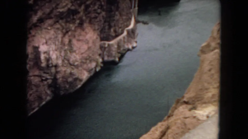 Visitor-shot 8mm frame of a rocky canyon wall, green-gray river, and a concrete chute-like dam feature descending near the water.