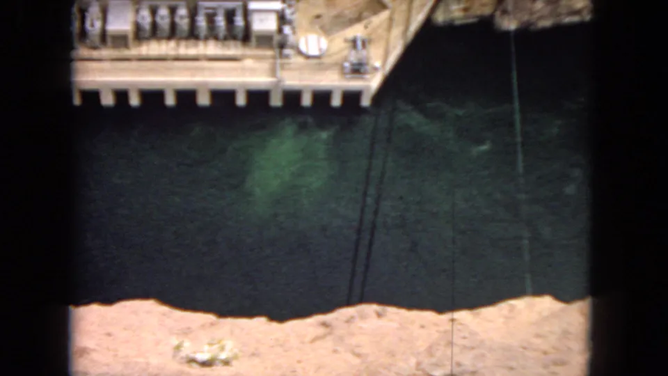 1967 Arizona frame showing part of a concrete dam or observation platform over dark river water within a desert canyon setting.