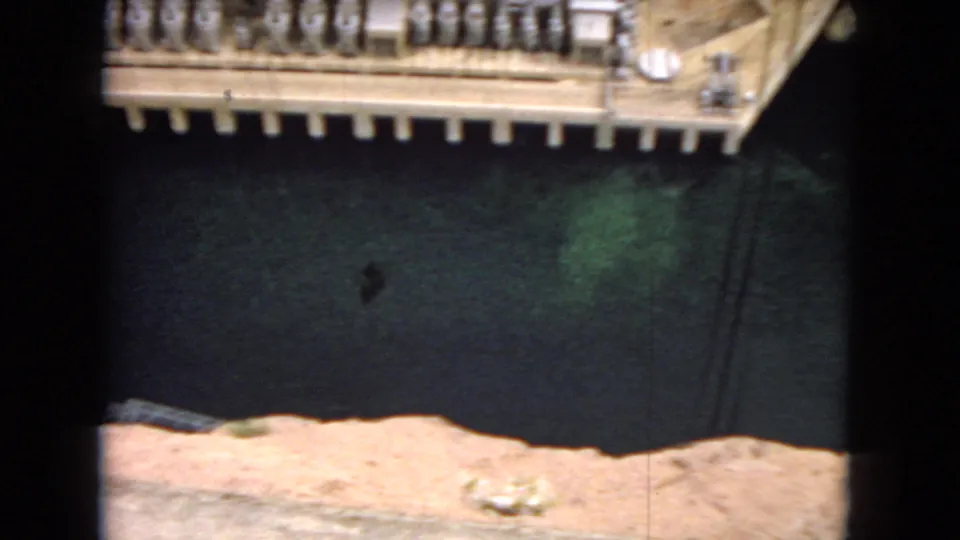 1967 8mm frame showing part of a hydroelectric dam model with a dark reservoir area, concrete structures, and rocky foreground terrain.