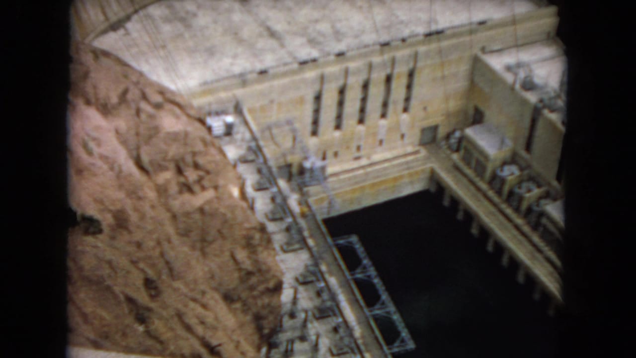 Visitor-shot 8mm view of a hydroelectric dam model, with a curved concrete dam face, rocky canyon sides, dark water, railings, and small equipment details.