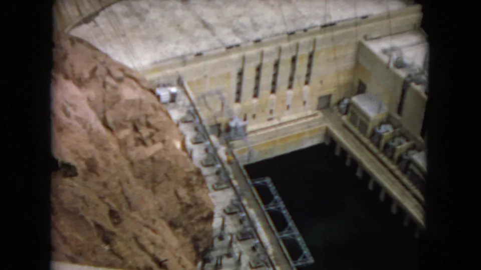Visitor-shot 8mm view of a hydroelectric dam model, with a curved concrete dam face, rocky canyon sides, dark water, railings, and small equipment details.