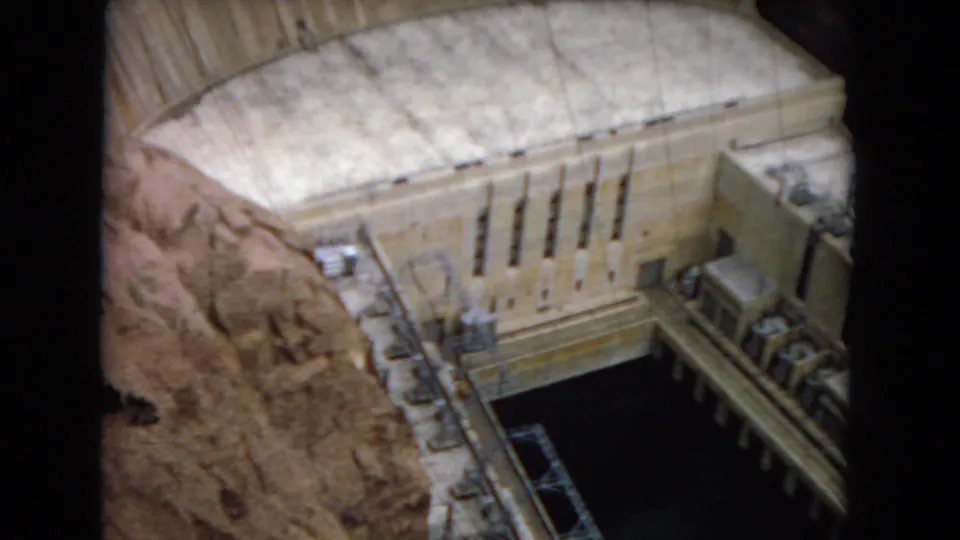 1967 home-movie frame showing a detailed hydroelectric dam model from above, including canyon rock texture, concrete openings, railings, and water-facing structures.