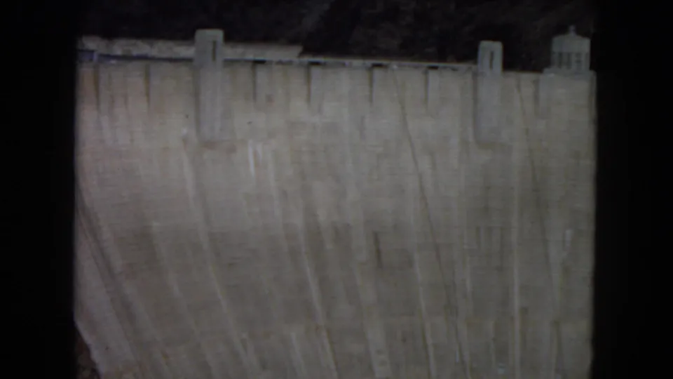 Wider 1967 8mm view showing a large concrete dam wall, vertical buttress-like forms, rocky desert terrain behind it, and visible film gate edges.