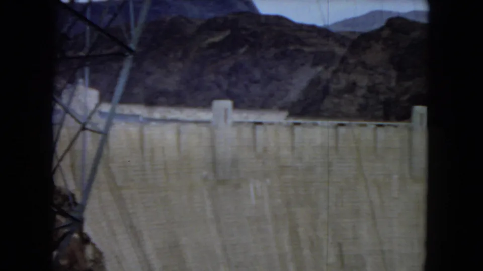1967 8mm view of a large concrete dam in an Arizona desert setting, with a steel transmission tower at left, rugged mountains behind, and a pale sky above.