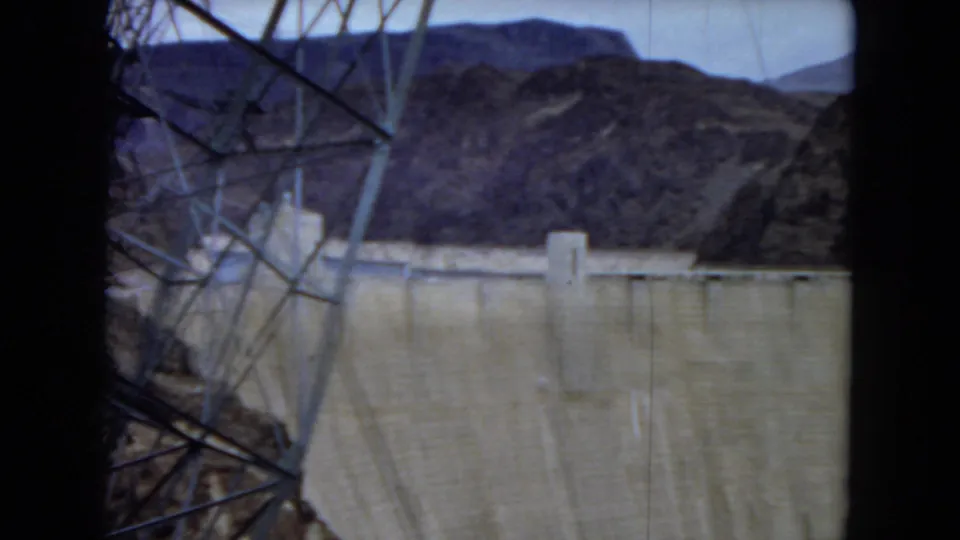 1967 8mm view of Hoover Dam through a dark window or structural frame, with a steel transmission tower in the foreground and desert mountains beyond.