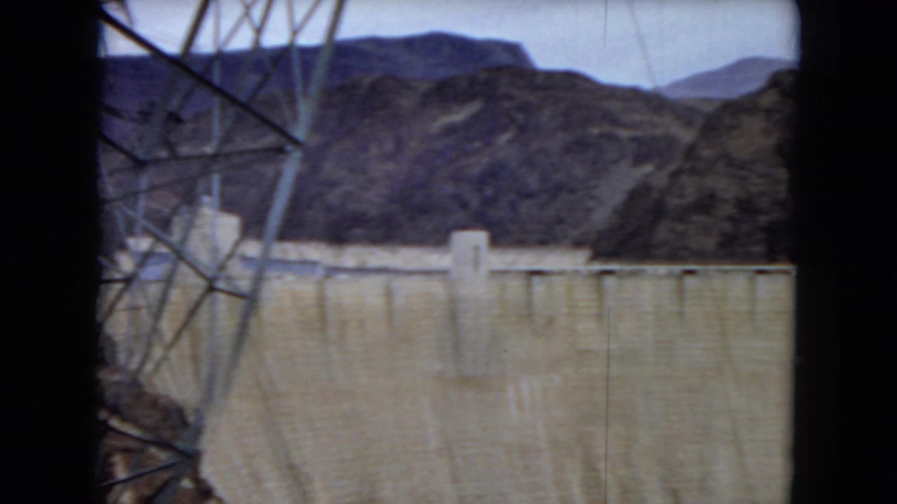 Hoover Dam appears beyond dark window framing, with the concrete wall, rugged hills, pale sky, and part of an electrical tower visible in the foreground.