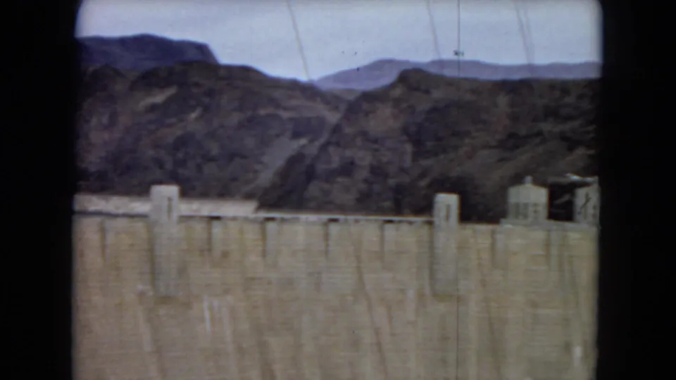 A visitor-shot 8mm frame shows the upper edge of Hoover Dam, desert mountains, pale sky, and a small structure near the dam edge.