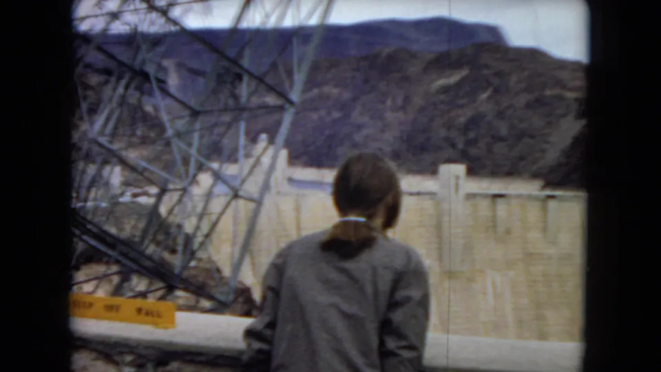 A woman in a dark jacket stands with her back to the camera, looking toward Hoover Dam through a framed opening with steel tower elements and desert mountains visible.