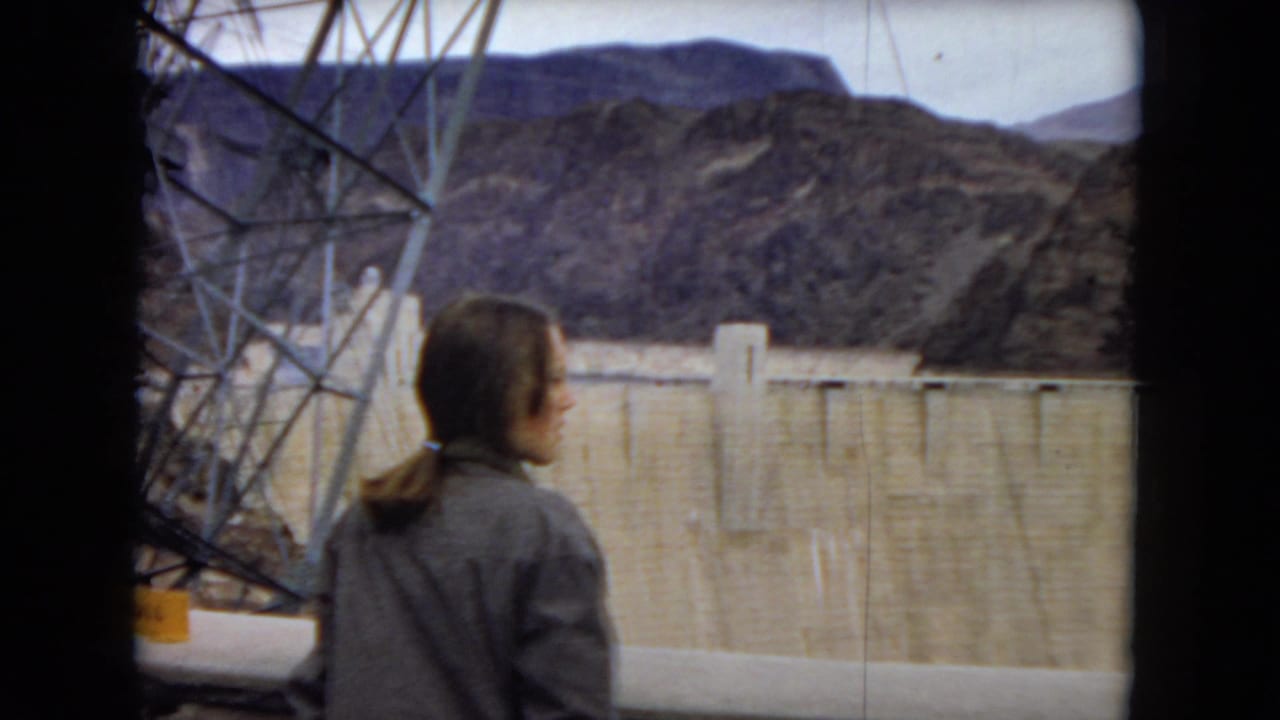 A woman in a jacket stands with her back to the camera near Hoover Dam, with steel framing, the concrete dam wall, and rocky mountains visible in the 1967 home-movie frame.