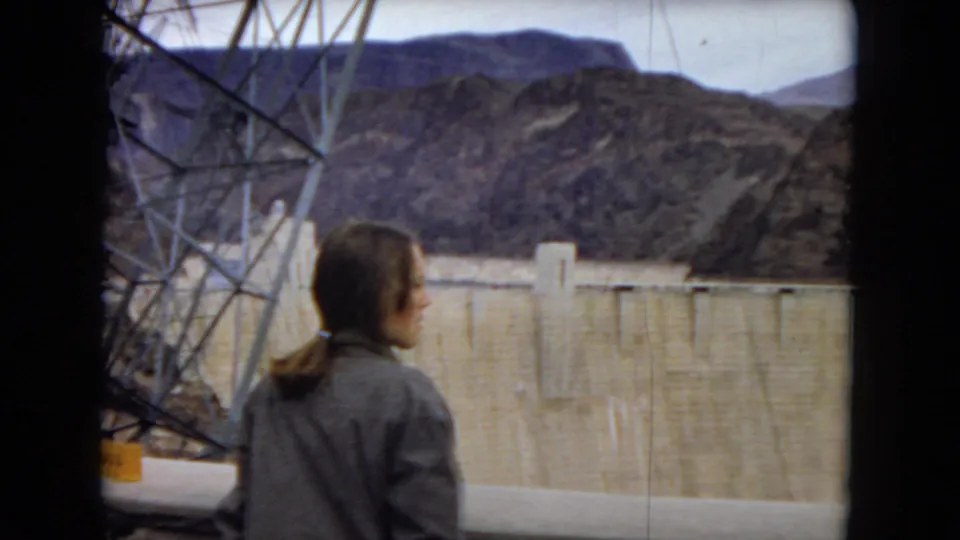 A woman in a jacket stands with her back to the camera near Hoover Dam, with steel framing, the concrete dam wall, and rocky mountains visible in the 1967 home-movie frame.