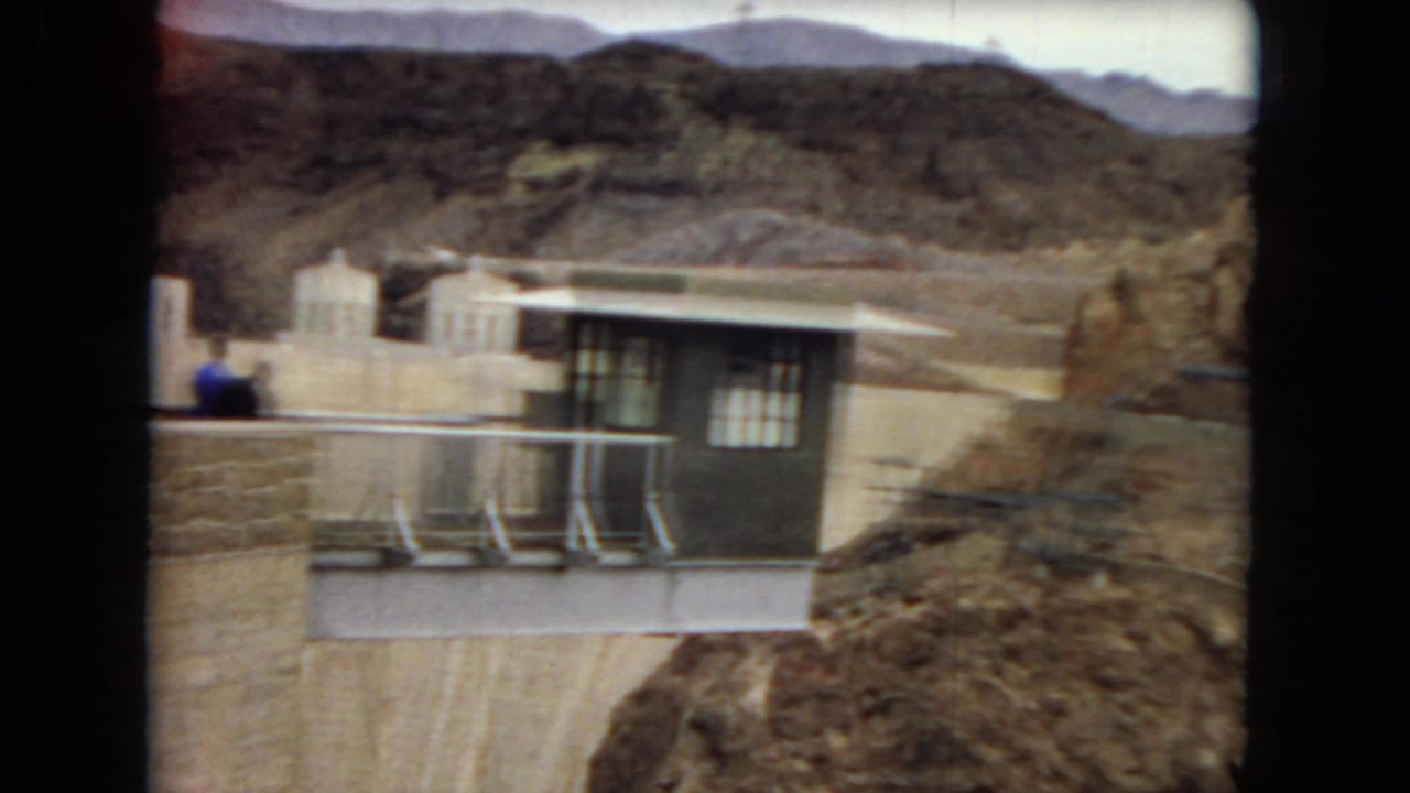 1967 8mm Arizona home-movie frame showing a dam-side control building, metal railings, rocky desert hills, and small distant visitors.