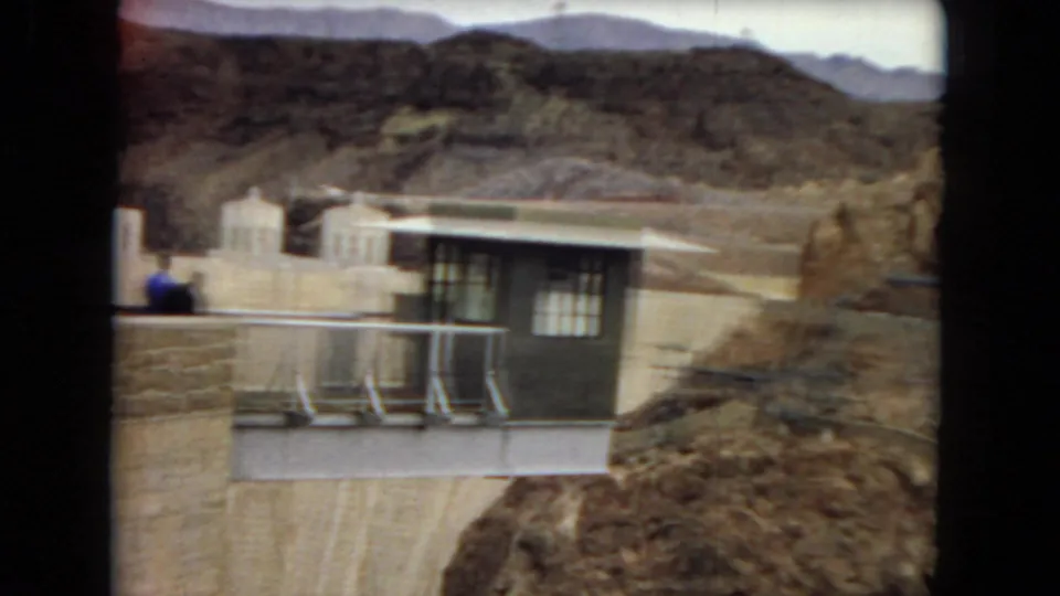 1967 8mm Arizona home-movie frame showing a dam-side control building, metal railings, rocky desert hills, and small distant visitors.