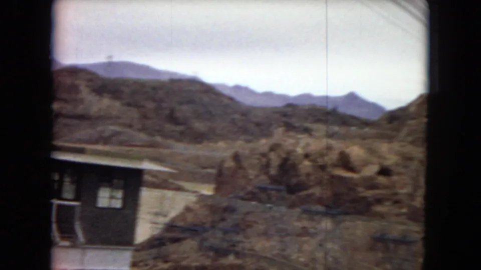 1967 8mm Arizona home-movie frame with the edge of a dam-side building, pale railings, arid rock formations, and mountains in the distance.