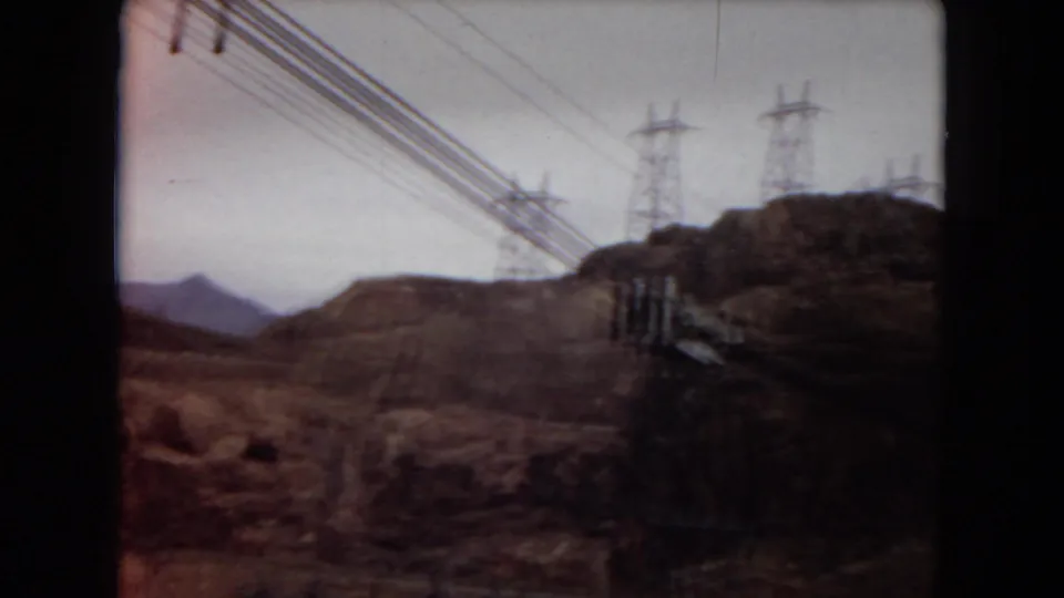 1967 Arizona 8mm frame showing overhead powerlines, metal transmission towers, rocky hills, and dry terrain beneath a pale sky.