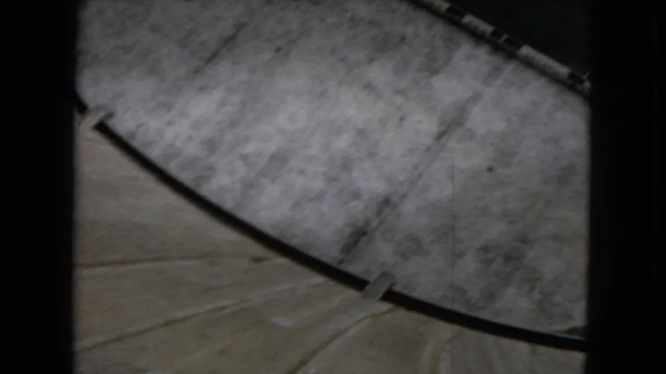 Downward 8mm view from the dam area, with pale concrete, a dark curved railing or cable, and gray paved surface visible below.