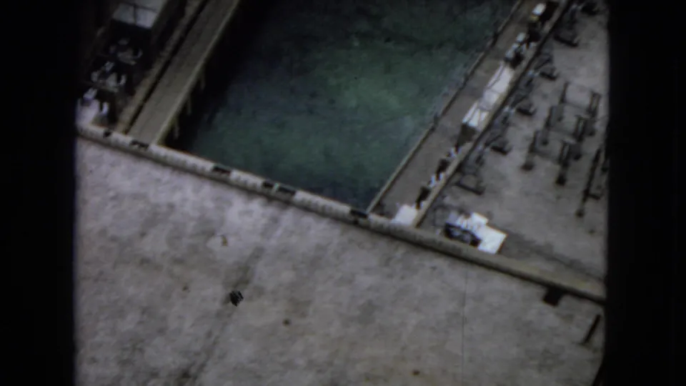 1967 8mm frame from an elevated dam-top viewpoint showing a rectangular water area, concrete deck, edge structures, and small equipment-like details below.