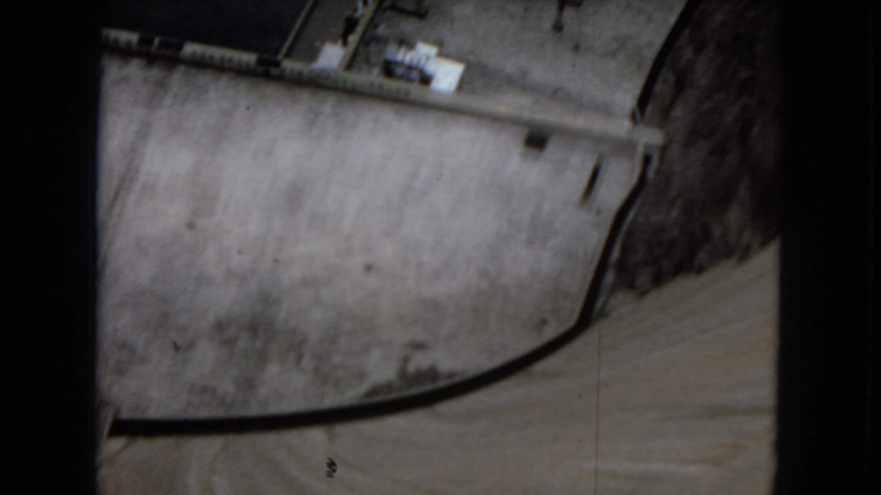 Overhead 1967 home-movie view of Hoover Dam concrete surfaces, showing a curved edge or roadway line and small structural details near the top of the frame.