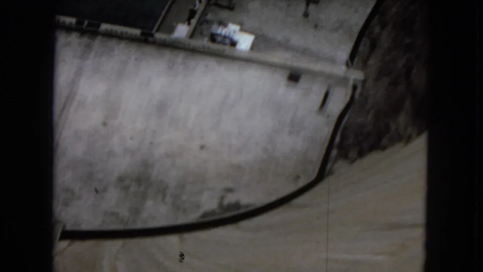 Overhead 1967 home-movie view of Hoover Dam concrete surfaces, showing a curved edge or roadway line and small structural details near the top of the frame.