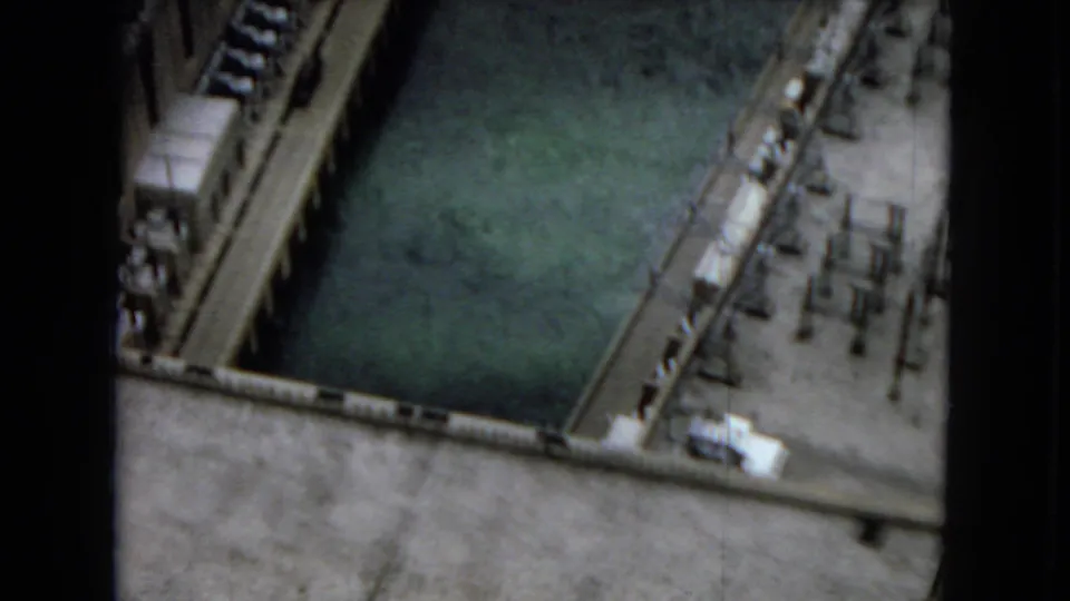 1967 8mm overhead view at Hoover Dam, looking down toward green-blue reservoir water bordered by concrete walkways, railings, and structural equipment.