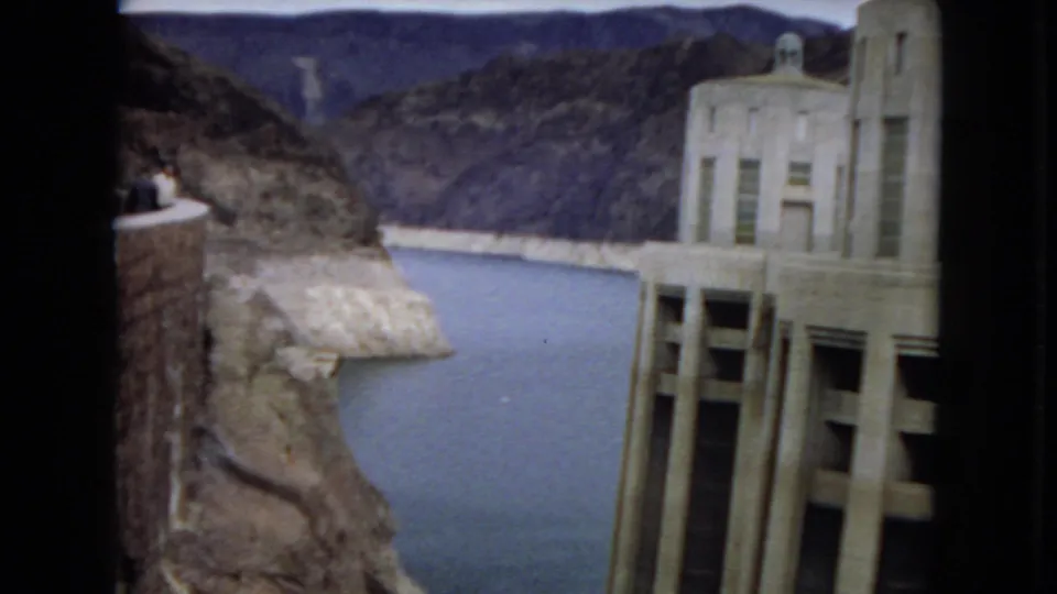 1967 8mm view of Hoover Dam with a concrete tower at right, reservoir water below, rocky canyon walls beyond, and a small overlook area at left.