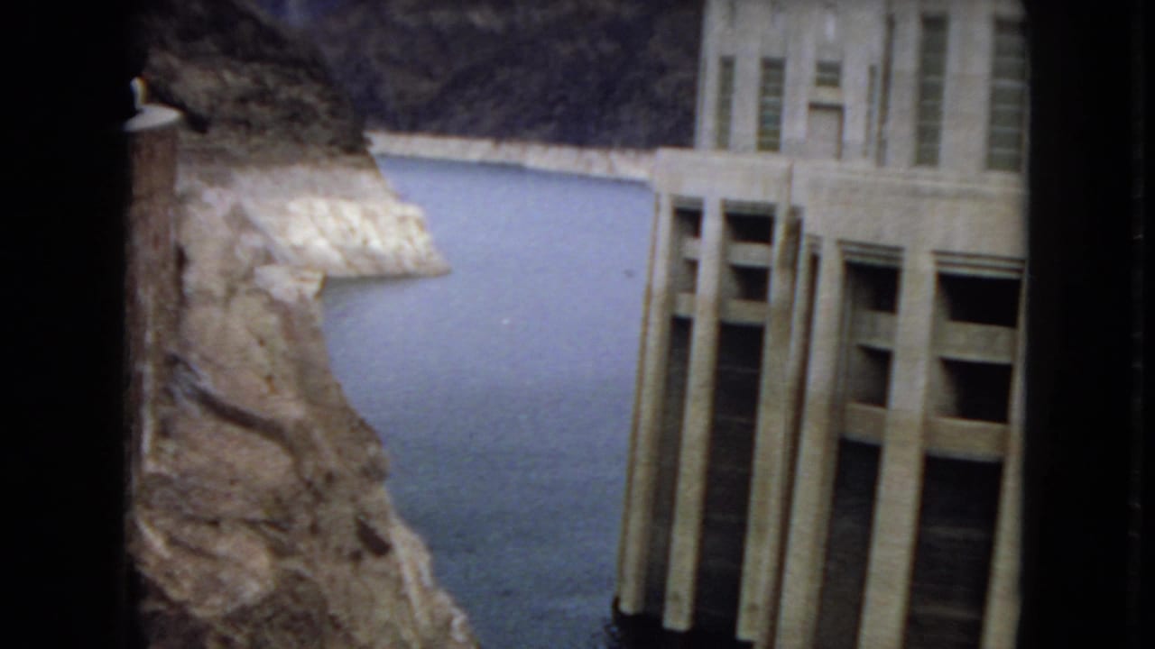1967 Arizona 8mm frame showing a tall concrete dam structure beside calm reservoir water and steep rocky cliffs.