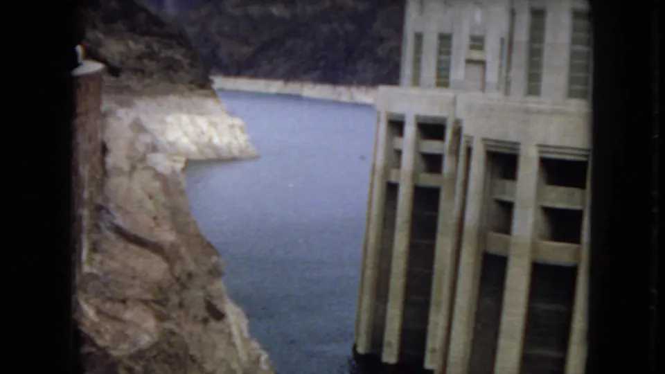 1967 Arizona 8mm frame showing a tall concrete dam structure beside calm reservoir water and steep rocky cliffs.