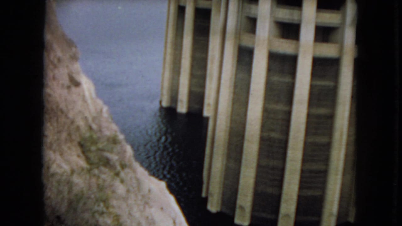 Close 1967 Arizona dam view with vertical concrete faces, dark waterline openings, still reservoir water, and a rocky slope at center.