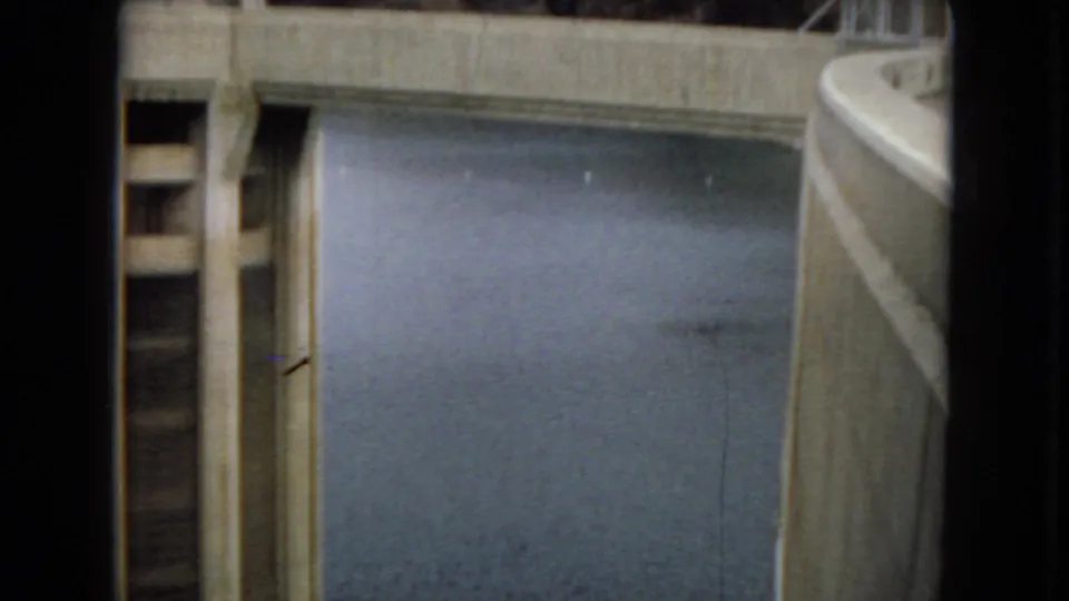 1967 Arizona frame showing a concrete bridge or deck crossing over calm water between large dam structures.