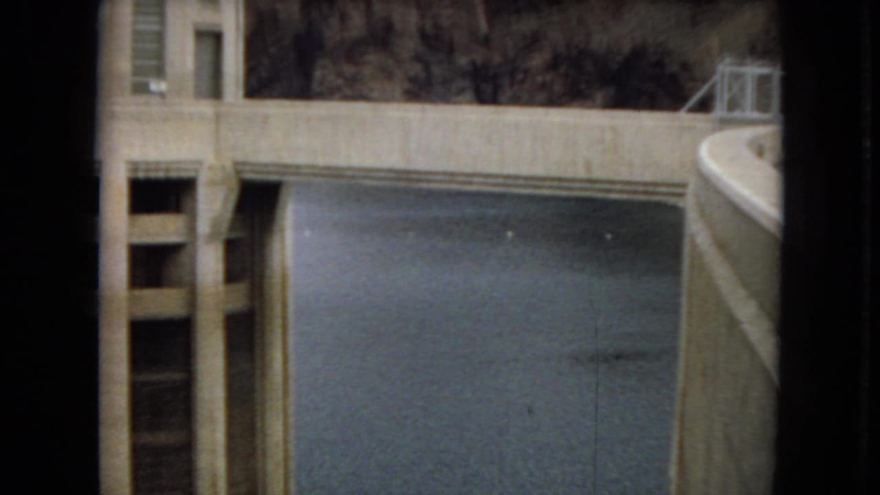 1967 home-movie frame showing a cropped view of Hoover Dam concrete structures, water, and a nearby utility building edge.