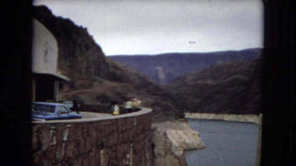 Visitor-shot 1967 frame at Hoover Dam showing a curving overlook road, parked period car, distant people, water below, and canyon slopes.