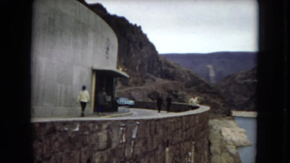 1967 home-movie view of visitors beside a concrete dam building, with a stone overlook wall, reservoir water, and rugged canyon terrain visible beyond.
