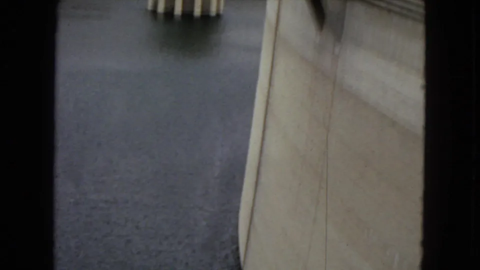 The camera frames the dam from very near the concrete face, emphasizing the reservoir surface, vertical wall, and overhead supports.