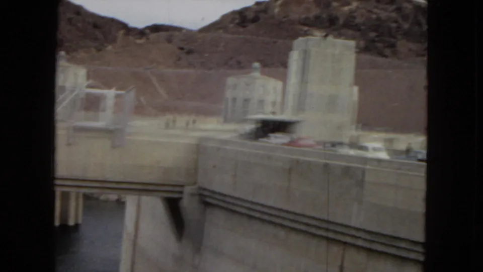 Hoover Dam roadway and concrete utility structures appear beside water and rocky desert slopes in a handheld 1967 8mm frame.