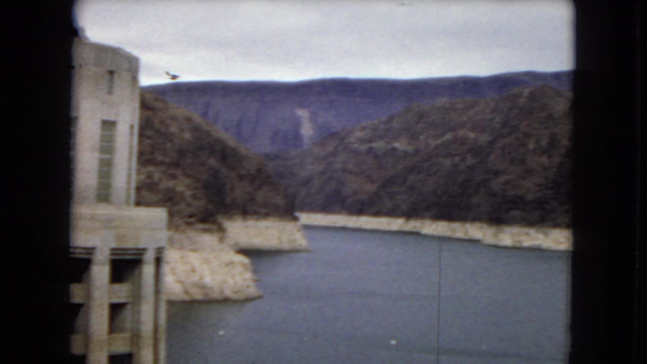 1967 8mm home-movie frame showing a concrete dam-side building edge, reservoir water, pale shoreline, and rugged desert canyon slopes.