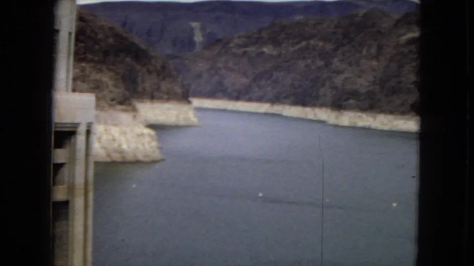 1967 8mm frame looking across reservoir water and pale canyon banks near the dam, with rugged desert mountains in the background.