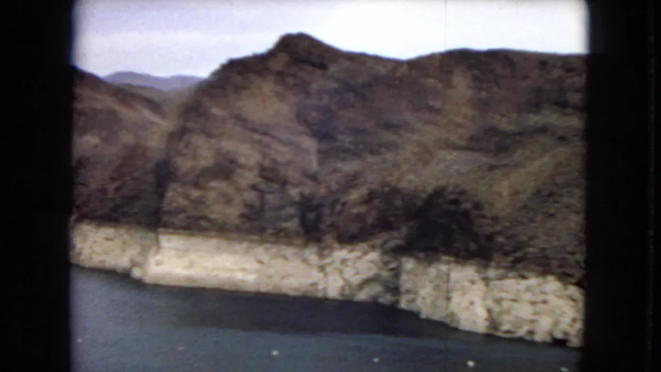 1967 8mm home-movie frame showing rugged canyon mountains, a pale exposed reservoir shoreline, and dark water below.