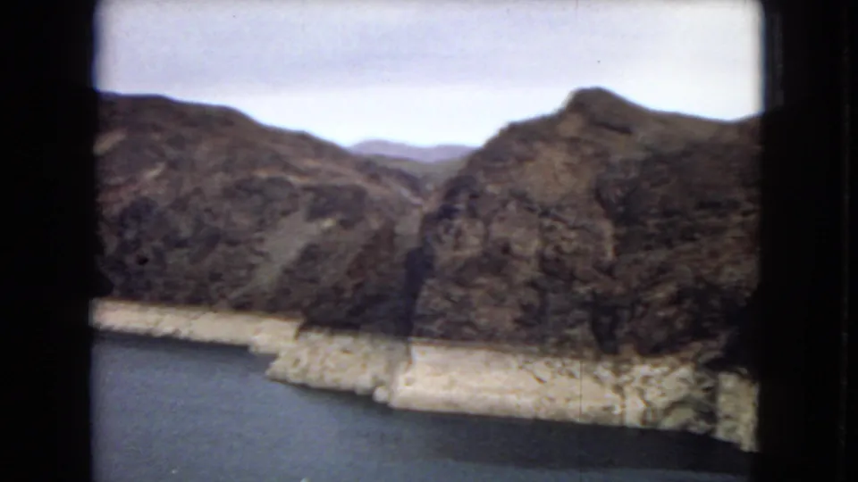 1967 8mm frame showing reservoir water below steep desert canyon ridges, with pale shoreline bands and soft handheld framing.