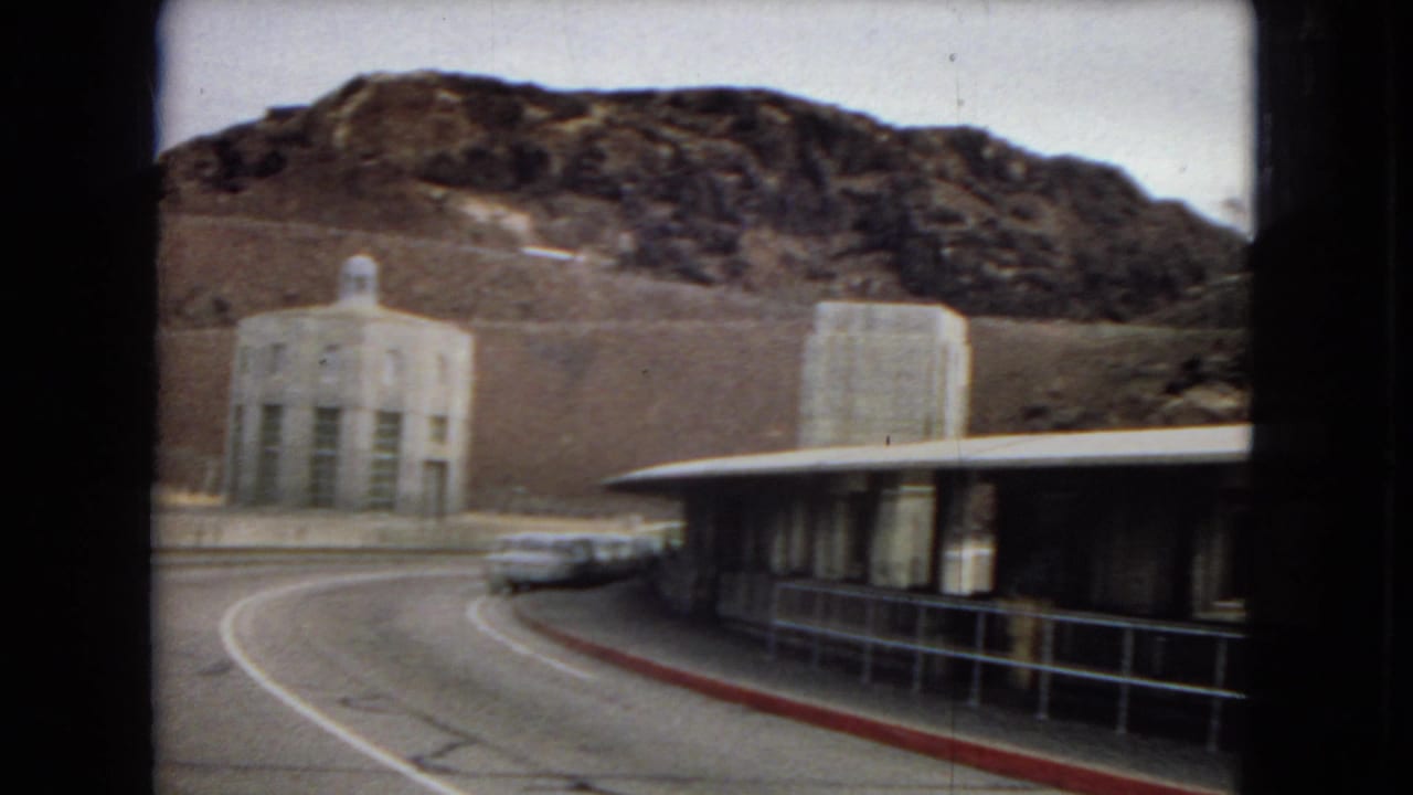 1967 8mm home-movie frame showing a curving road, a white station wagon, a long low building, and a stone tower against rugged desert hills near the Boulder Dam area.