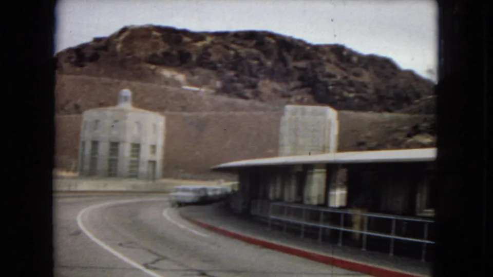 1967 8mm home-movie frame showing a curving road, a white station wagon, a long low building, and a stone tower against rugged desert hills near the Boulder Dam area.