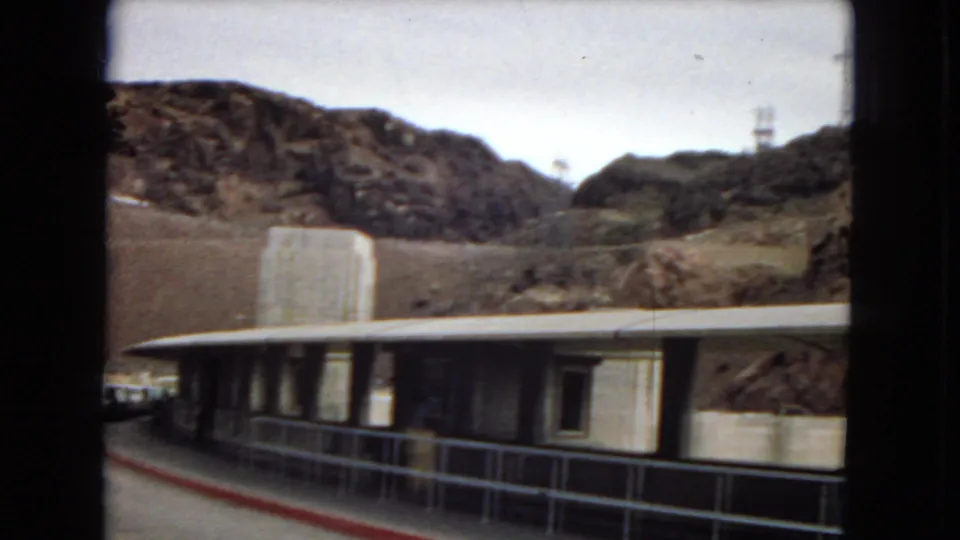 Vintage 8mm frame of a low roadside structure with a railing and stone tower, backed by rocky desert terrain and a visible utility pole.