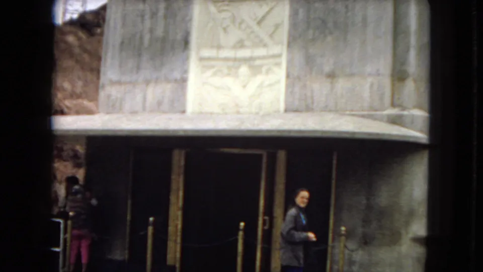 Home-movie frame showing a stone entrance with carved relief panels above the doorway, with visitors visible near the roped entry area.