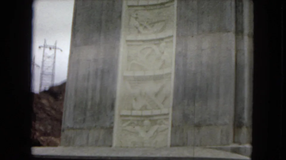 Close 8mm view of carved relief panels set into a tall stone wall, with desert hillside and utility poles visible at the side of the frame.