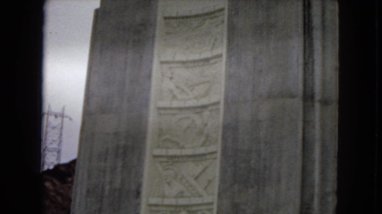 1967 Arizona home-movie frame showing the upper portion of a massive concrete dam structure with vertical decorative relief panels and dark film-edge borders.