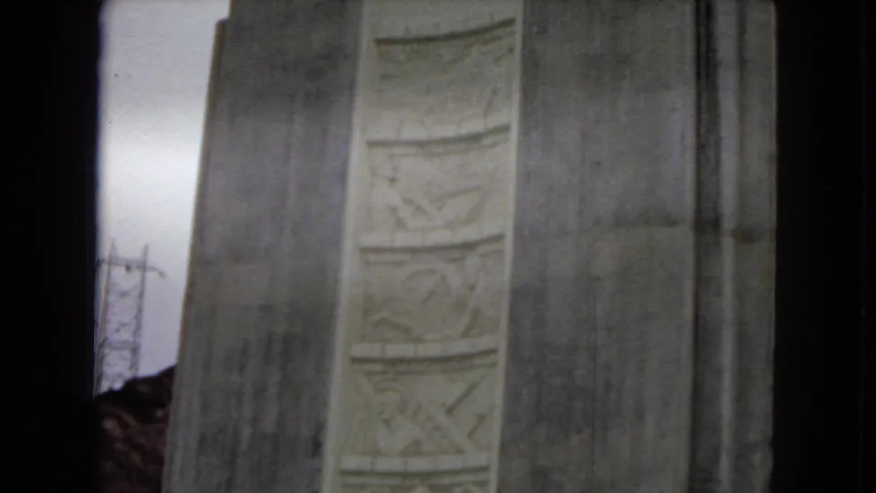 1967 Arizona home-movie frame showing the upper portion of a massive concrete dam structure with vertical decorative relief panels and dark film-edge borders.