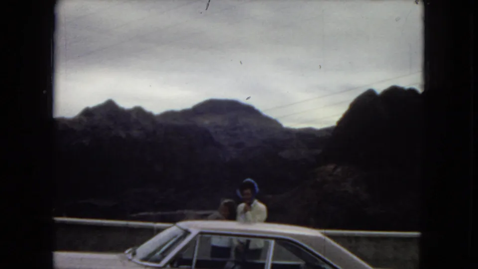 1967 Arizona home-movie frame showing a woman and smaller companion standing by a classic car at a roadside viewpoint with mountains behind them.