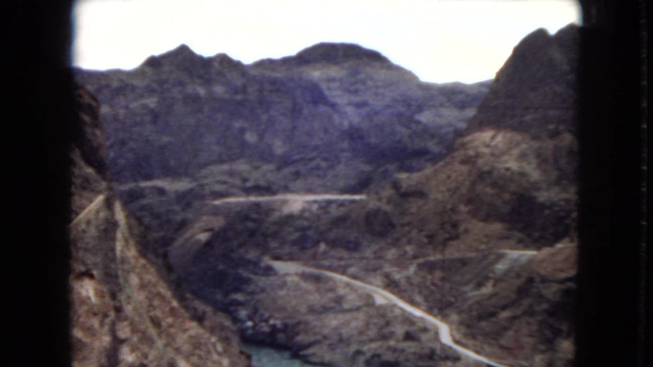 1967 8mm frame from Arizona showing rugged canyon mountains, a pale winding road, and a small stretch of water below an overexposed sky.