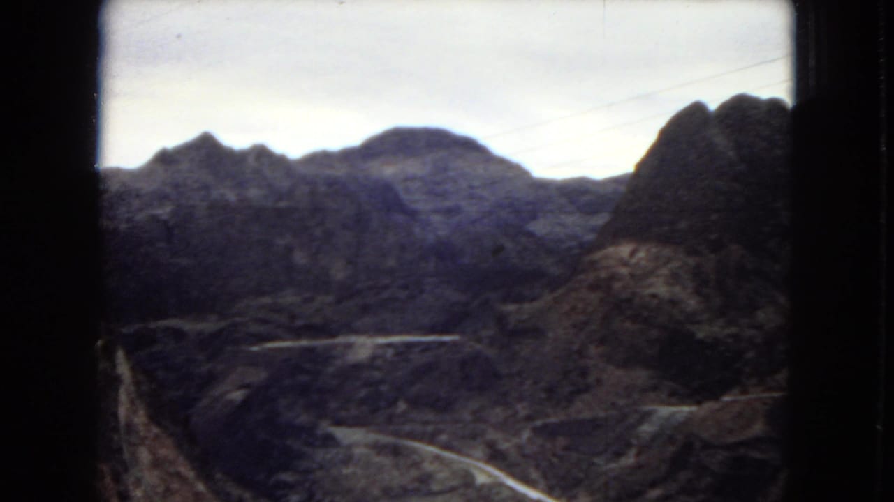 1967 Arizona home-movie frame with dark 8mm frame borders, rocky ridges, a winding road, and canyon terrain in soft faded color.